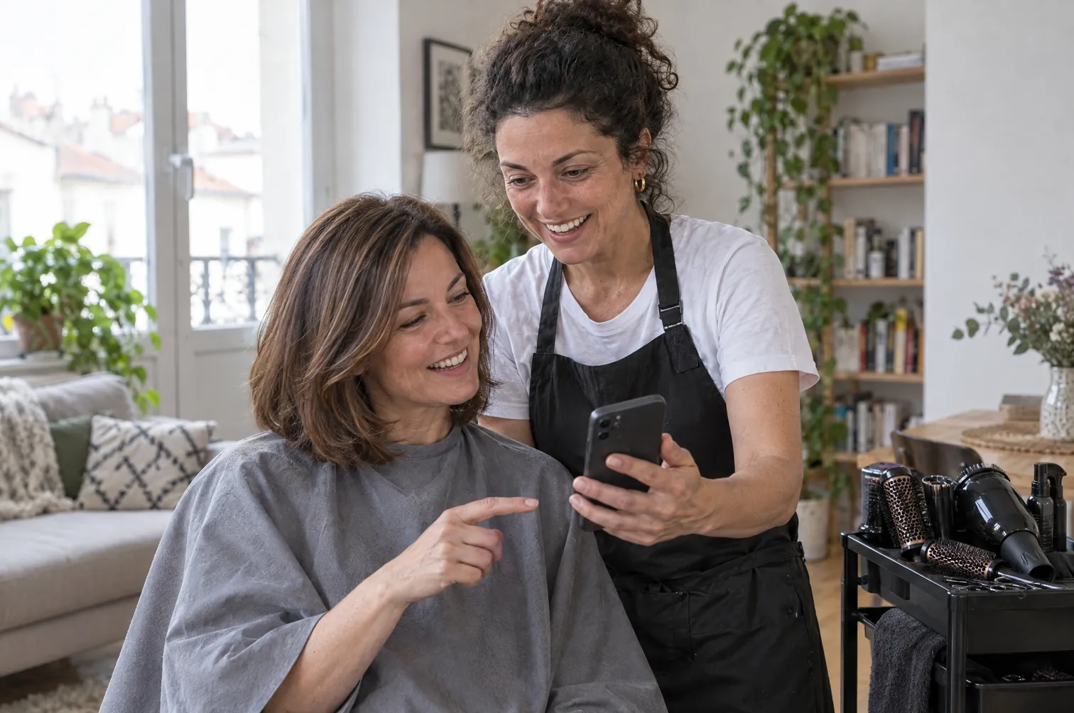 Une coiffeuse à domicile montre sa nouvelle page internet sur son téléphone à une cliente, dans le salon de celle-ci